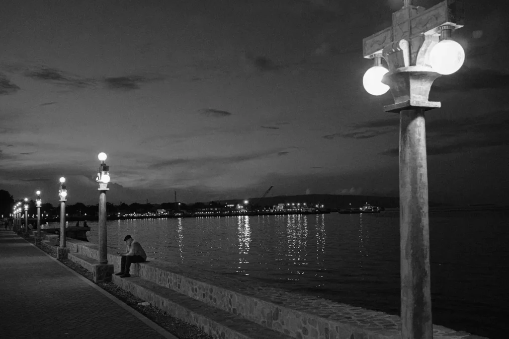a black and white photo of a lone man sitting on dumaguete boulevard next to the sea. Photo taken on the Fuji X100VI