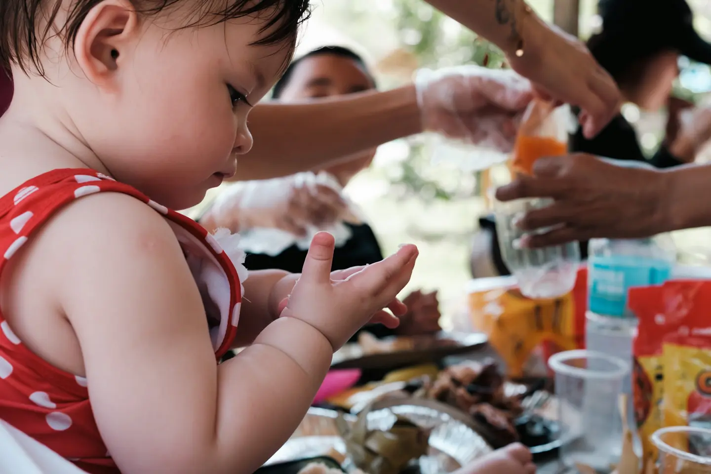 a young girl stares at rice sticking to her fingers while others reach for food and drink in the background.