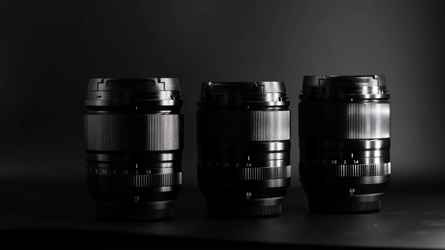 The Fuji 18mm 1.4WR lens next to the Fujifilm 23mm 1.4WR and Fuji 33mm 1.4WR on a black background in dramatic light.
