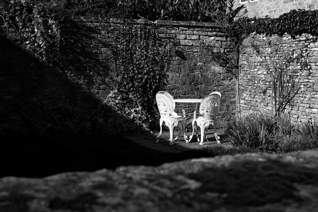shot of a back garden in the Cotswolds in black and white using the Fujifilm X-T5 camera. It shows a dark shaded area and a well lit area with ornamental garden table and chairs. 