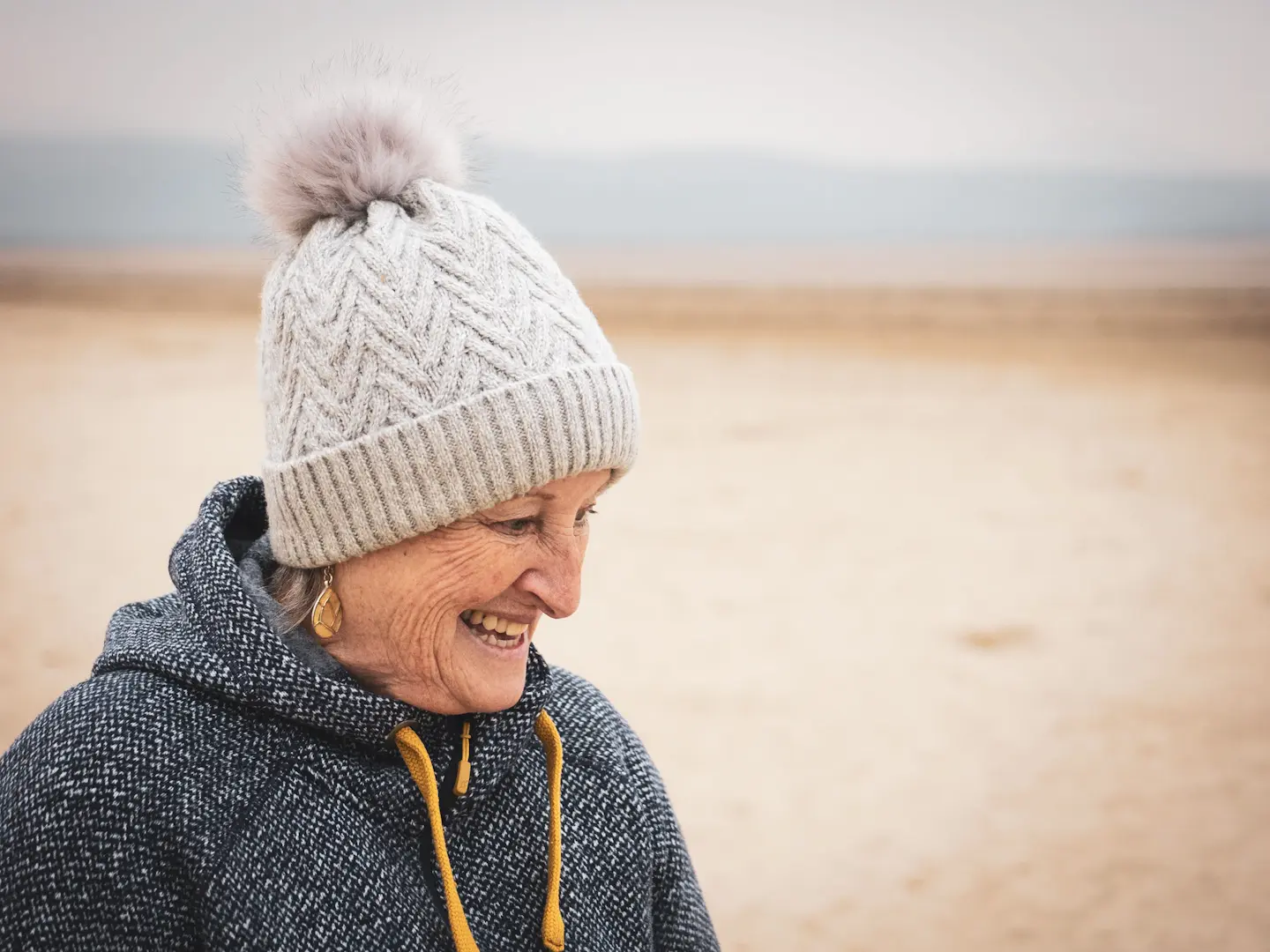 A simple portrait of an elderly lady at the beach in Wales with a bobble hat and fleece jacket on.