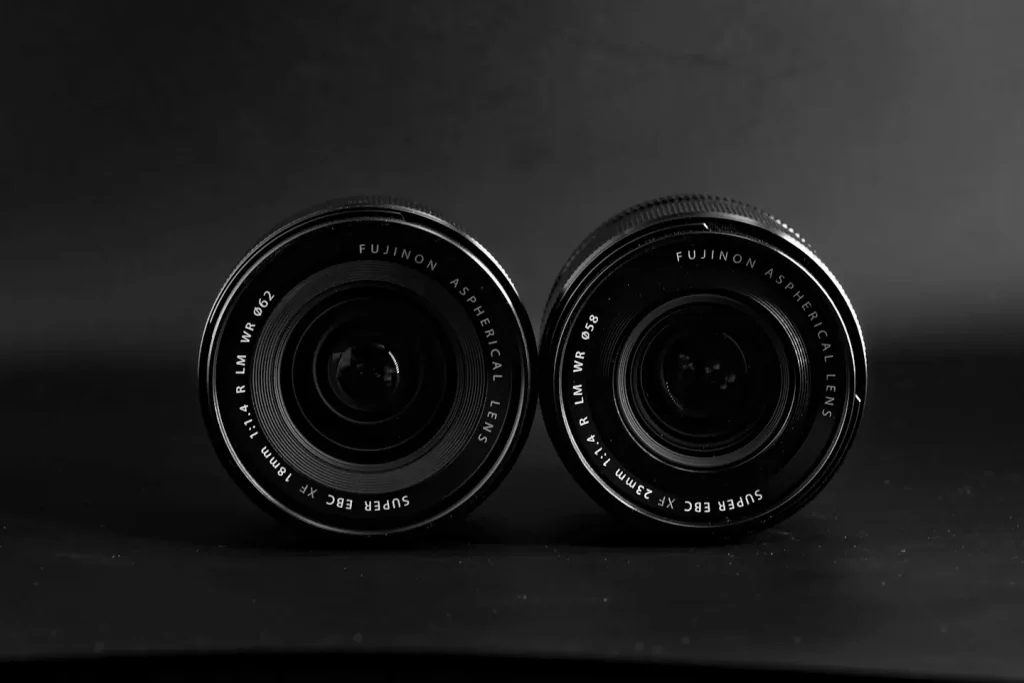 The front lens elements of the Fuji 18mm 1.4WR lens and the Fuji 23mm 1.4WR lens next to each other on a black background in dramatic lighting. 