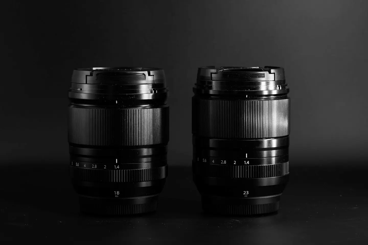 The Fujifilm 18mm 1.4WR lens next to the Fujifilm 23mm 1.4WR lens on a balck background in dramatic light.