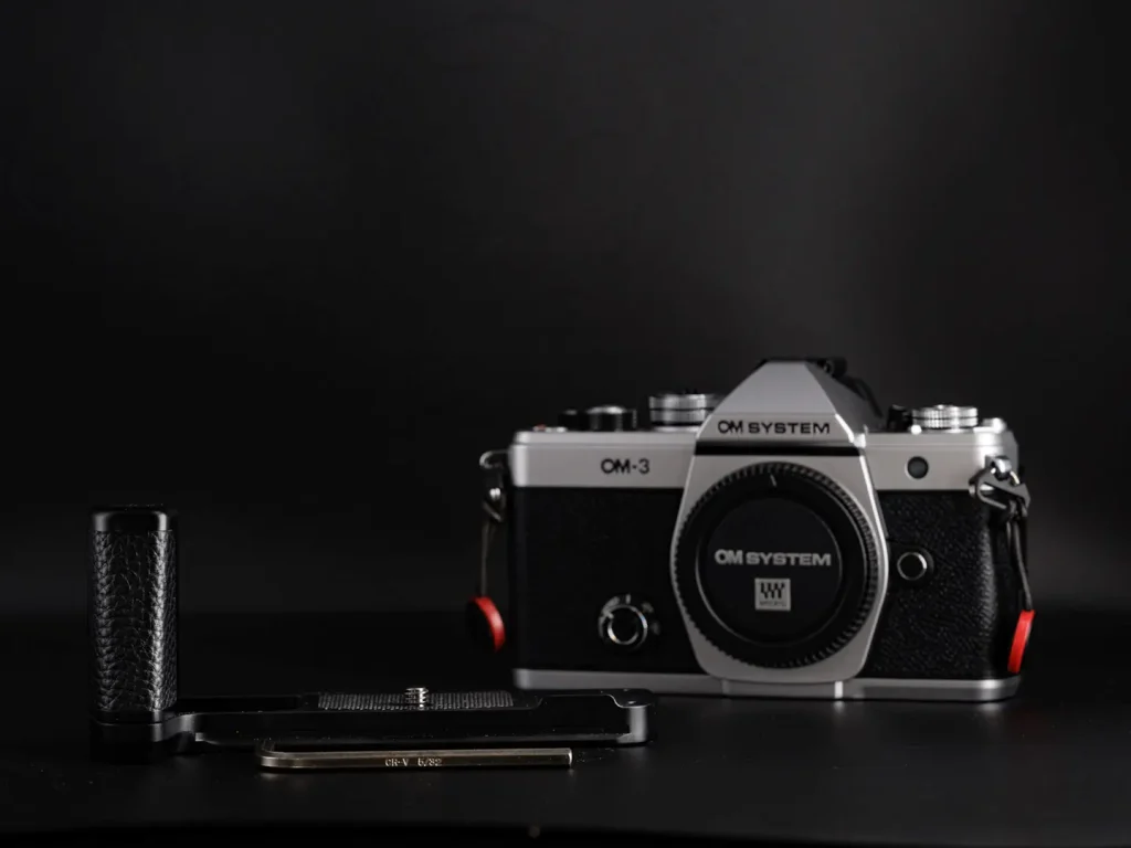 Haoge hand grip for the Om System OM-3 camera photographed next to the camera on a black background.