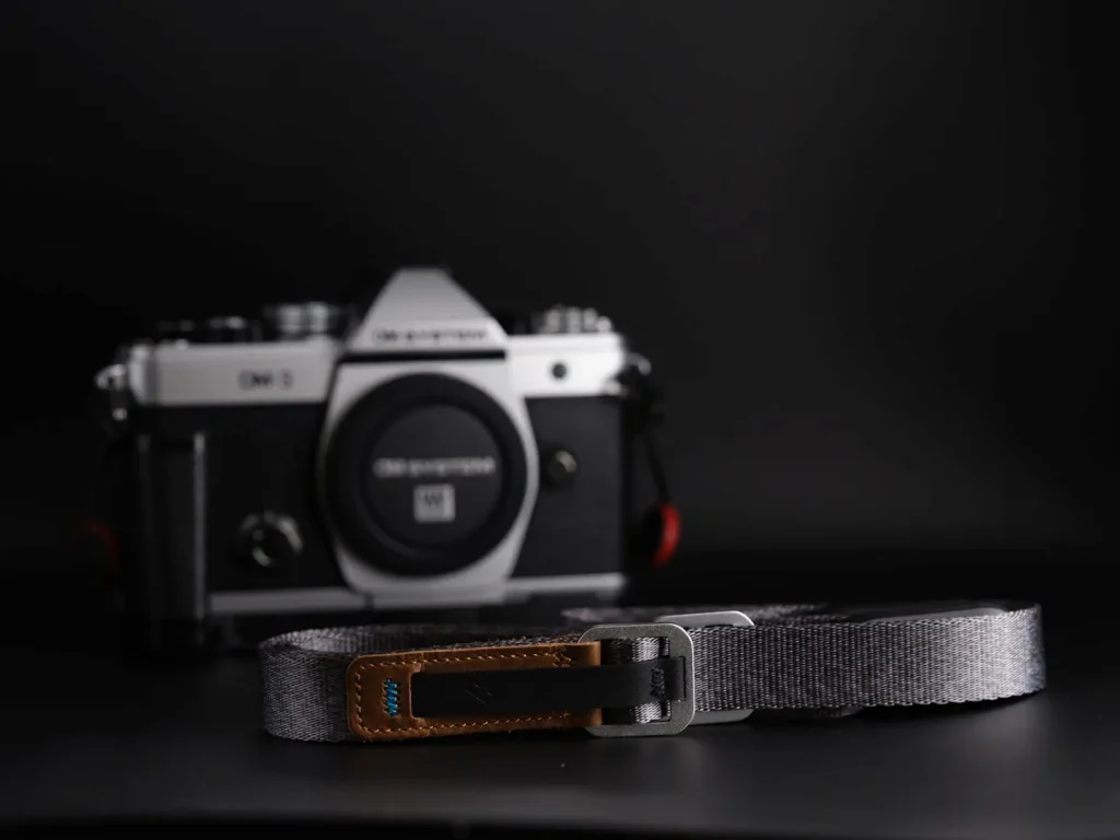 Peak Design camera leash camera strap photographed next to an OM System OM-3 camera on a black background.