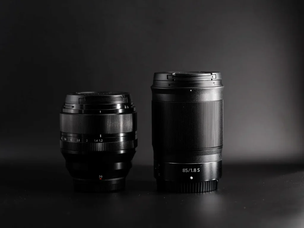 The Fujifilm XF 56mm 1.2WR lens next to the Nikon Z 85mm 1.8 lens to show the comparative size of each lens. Photo is taken on a black background.