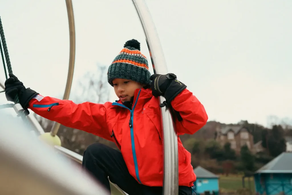 A 9 year old girl playing on a climbing frame at a park in Scotland, dressed in a red waterproof jacket, gloves and a hat. 