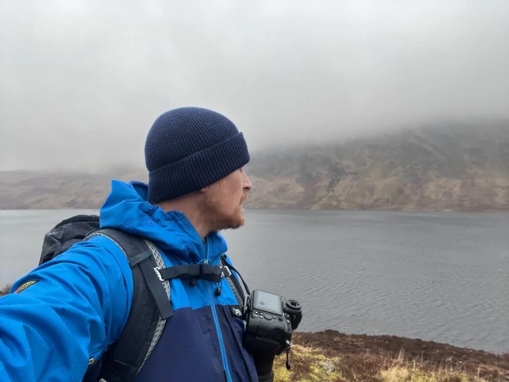A photographer stands in a Scottish glen in wind driven rain with his Nikon Z8 camera attached to his rucksack with a Peak Design Capture Clip.
