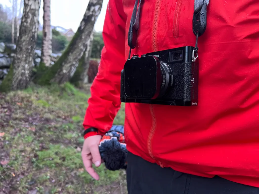 A photographer in a red waterproof jacket has the Fujifilm X100VI camera hanging around his neck on a Peak Design Camera Leash strap. 