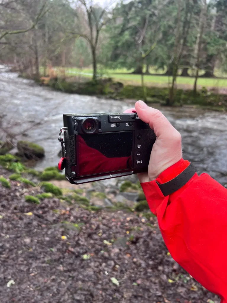 A photographer holds his Fuji X100VI out in front of him to frame a composition on the rear LCD screen by a Scottish river. 