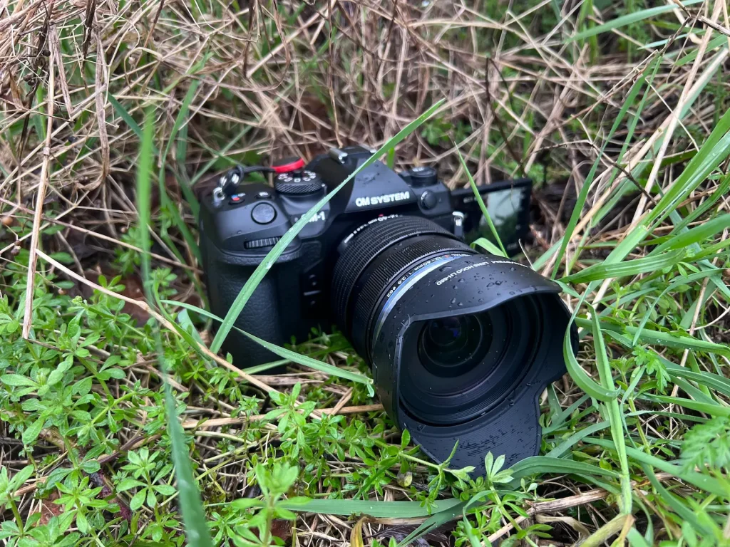The OM System OM1 Mark II camera sat in wet grass and twigs while covered in rain.