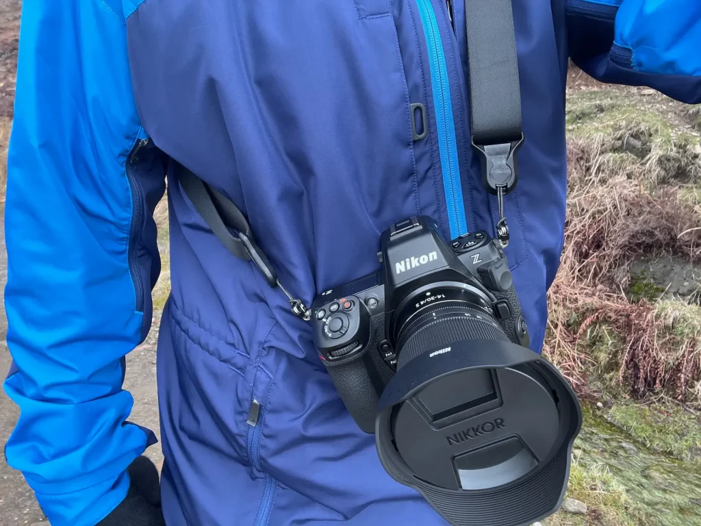 A Nikon Z8 + 14-30mm f/4 lens hanging from a photographers camera strap. The photographer is dressed for the outdoors in a Paramo Alta III jacket which is blue coloured. 