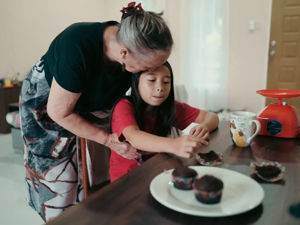 A grandma leans in to give her grandaughter a kiss from behind while the grandaughter is busy eating the muffins that they have just cooked together. Photo is taken on a Fujifilm GFX100S camera.
