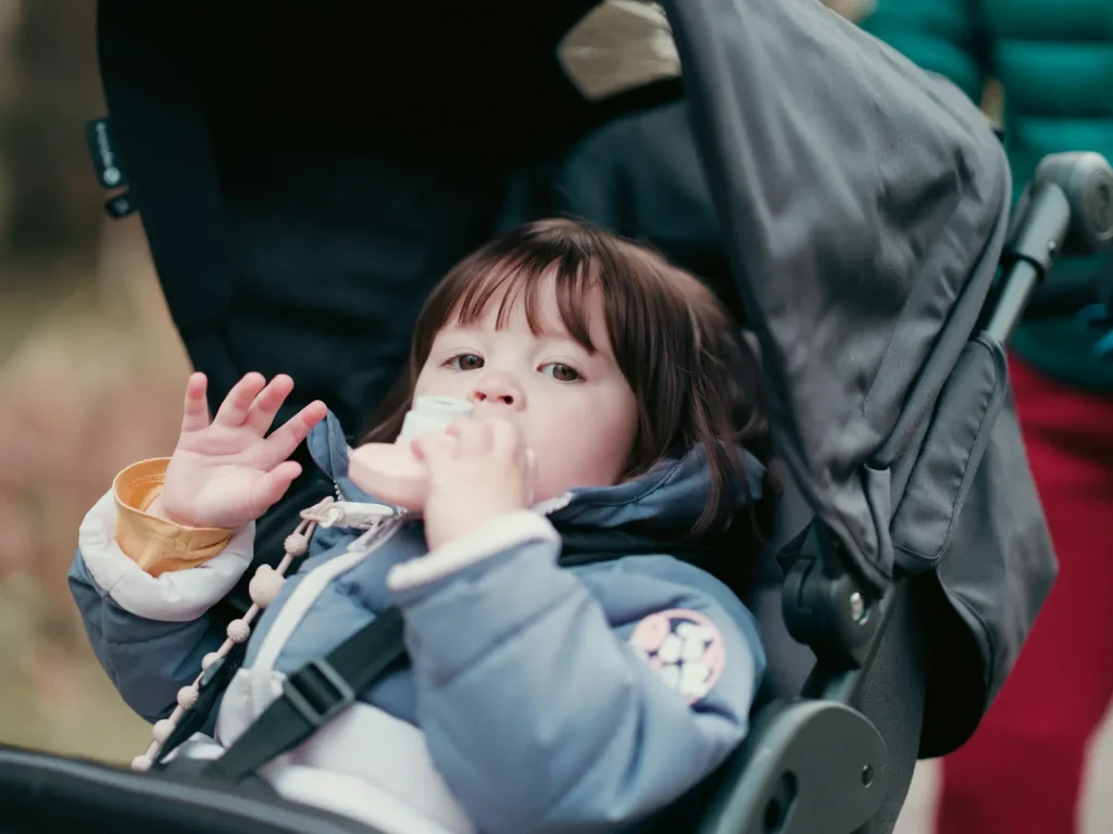 A young girl in a pram wearing a padded jacket while waving to the camera.