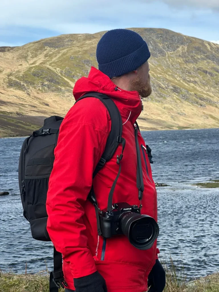 A photographer in a red waterproof jacket looks out on to Loch Turret while his Fuji GFX100S camera and GF lens are carried on his camera strap. 
