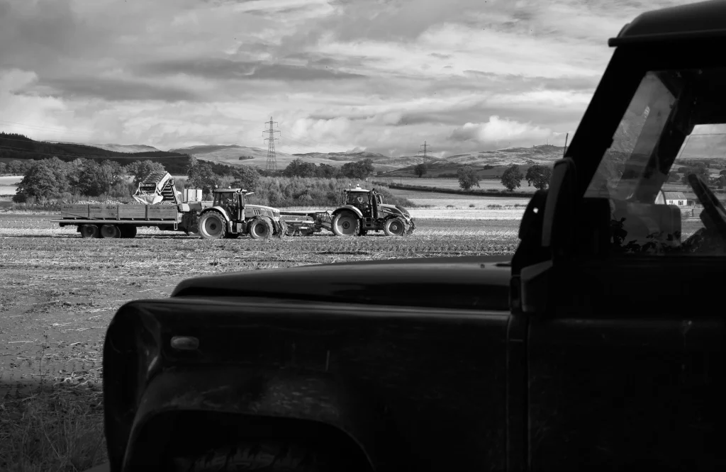 A black and white photo of two tractors in a field in Crieff, harvesting potatos. In the foreground is the outline of a Land Rover Defender.