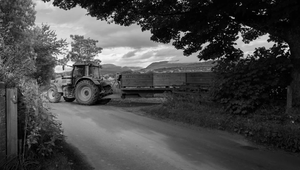 A black and white photo of a tractor pulling a truck-load of potatos that have just been harvested. The tractor is moving out of a filed on to a country lane.