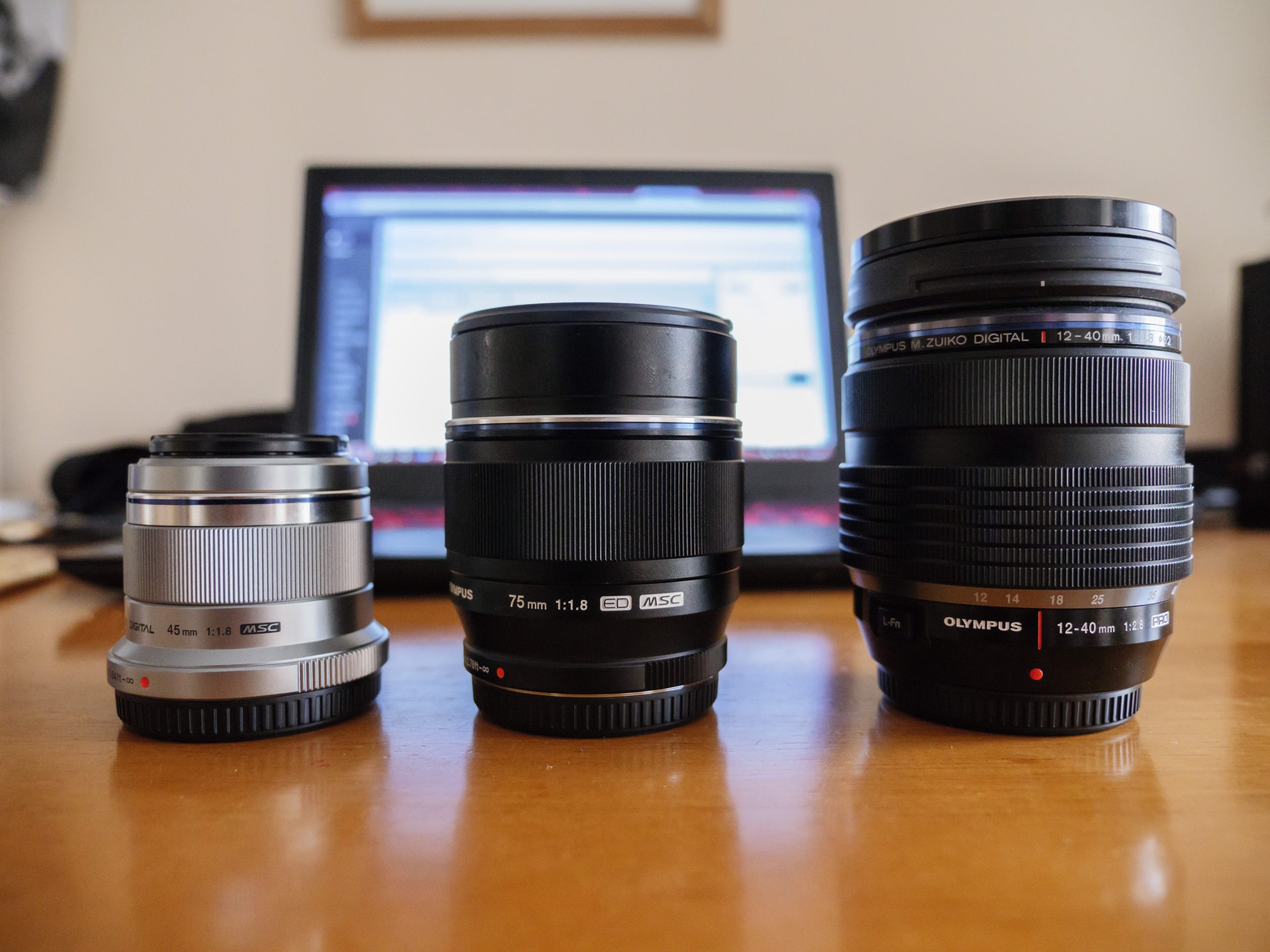 The Olympus 75mm 1.8 lens between the 45mm 1.8 and 12-40mm f/2.8 Pro lenses on a computer desk.
